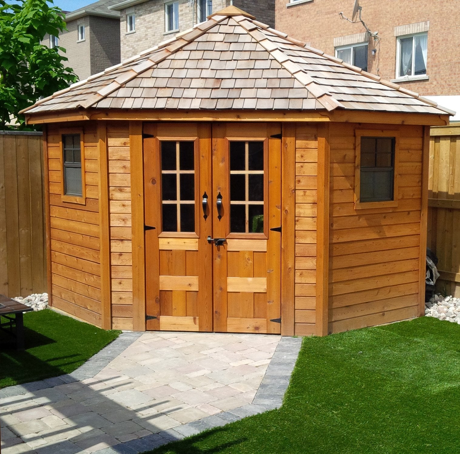 Front view of the Penthouse Garden Shed. The five sided shed is placed in the corner of a fenced backyard and concrete blocks pave the way into the shed.